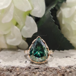 May include: A pear-shaped green gemstone ring with a gold setting and a delicate band of small diamonds. The ring is set against a background of white flowers.