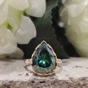 May include: A pear-shaped green moissanite engagement ring with a gold band and small diamonds on the band. The ring is set on a gray background.