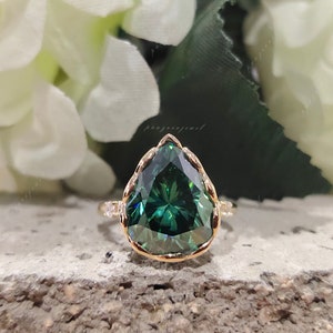 May include: A pear-shaped green gemstone ring with a gold band and a delicate, intricate design. The ring is set against a backdrop of white flowers.