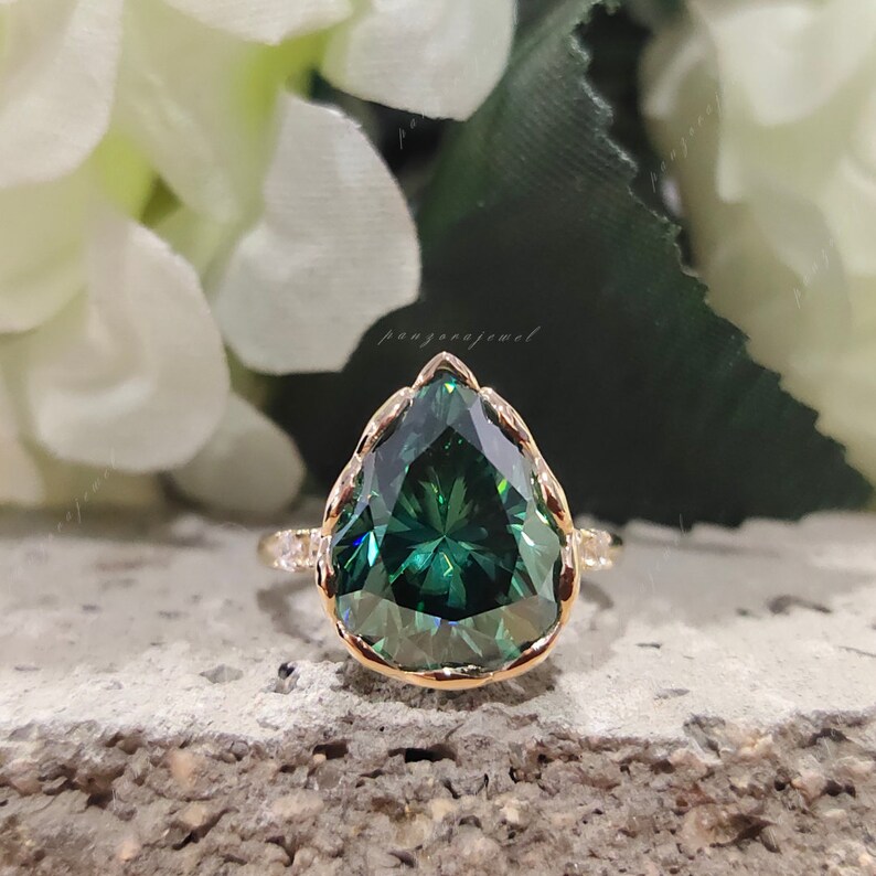 May include: A pear-shaped green gemstone ring with a gold band and a delicate, intricate design. The ring is set against a backdrop of white flowers.