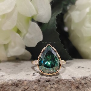 May include: A pear-shaped green gemstone ring with a gold band and a delicate, scalloped setting. The ring is set against a background of white flowers.