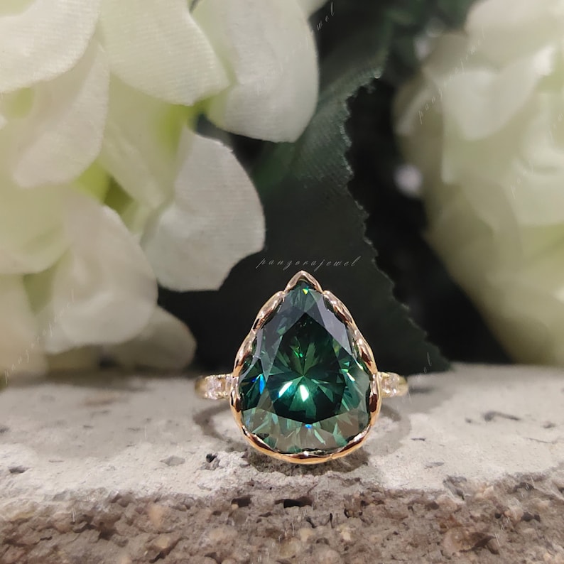 May include: A pear-shaped green gemstone ring with a gold band and a delicate, scalloped setting. The ring is set against a background of white flowers.