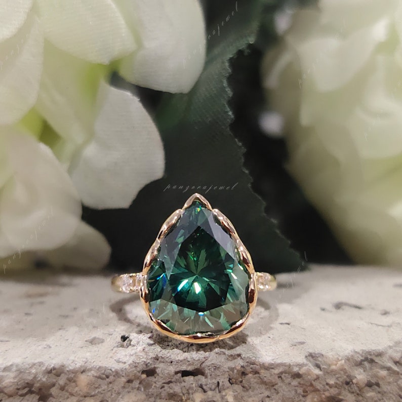May include: A pear-shaped green gemstone ring with a gold band and a delicate, scalloped setting. The ring is set against a backdrop of white flowers.