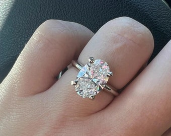May include: A silver ring featuring a large, oval-cut diamond. The diamond is the focal point, set with four prongs on a thin silver band. The ring is shown on a finger.