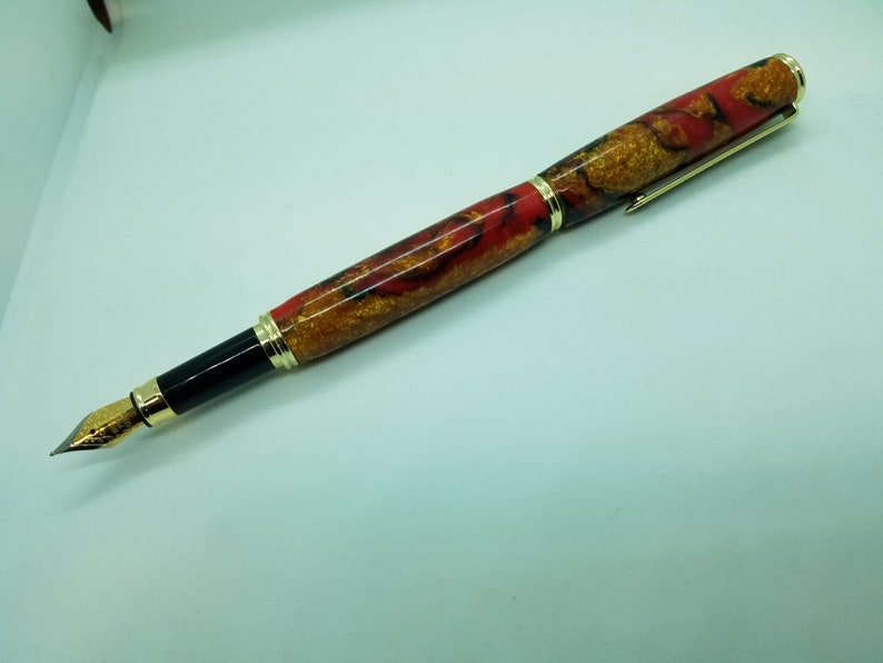 May include: A red and gold fountain pen with a black ink cartridge and a gold clip. The pen has a marbled design.