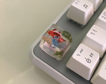 May include: A clear resin artisan keycap featuring an embedded miniature red and black koi fish, a small seashell, and green aquatic plant elements. The square keycap is positioned on a gray keyboard base next to white keys with black symbols.