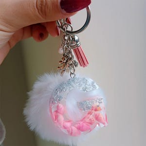 Op de afbeelding: Een handgemaakte sleutelhanger met een heldere harsletter "G" gevuld met roze schelpen en zilveren glitter. De sleutelhanger heeft een pluizige witte pompon, een roze kwast en zilveren bedels. De sleutelhanger is bevestigd aan een zilveren sleutelring.
