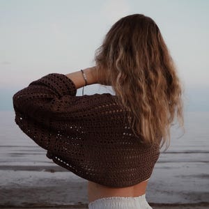 Könnte beinhalten: Eine Frau von hinten, die ein braunes Häkeltop mit langen Ärmeln trägt. Das Top ist offen gewebt und cropped. Die Frau hat lange, wellige blonde Haare. Der Hintergrund zeigt das Meer und den Himmel.