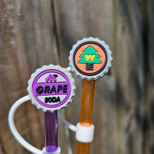 May include: Two colorful plastic straws with decorative tops. One straw has a purple top with the words "Grape Soda" and a cluster of grapes. The other straw has an orange top with a green pine tree and the letter "W" inside a circle.
