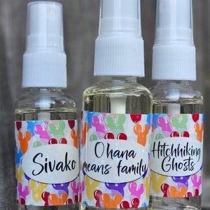 Puede incluir: Tres botellas de spray de vidrio transparente con tapas blancas y etiquetas coloridas. Las etiquetas presentan orejas de Mickey Mouse y globos. El texto en las etiquetas dice "Sivako", "Ohana means family" y "Hitchhiking Ghosts".