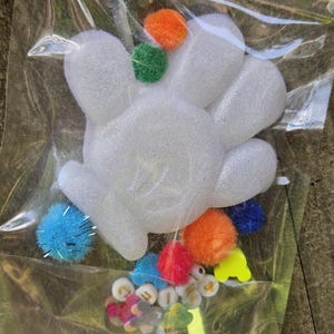 May include: A white, hand-shaped craft foam piece with colorful pom-poms and small decorative embellishments inside a clear plastic bag. The pom-poms are in various colors, including orange, green, blue, and red. The embellishments include small flower and letter-shaped pieces.