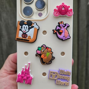 Könnte beinhalten: Eine weiße Handyhülle mit verschiedenen bunten Disney-Charakter-Anhängern. Die Anhänger sind Mickey Mouse, ein Drache, ein Schloss, eine Kutsche und ein Walt Disney World-Logo. Die Hülle hat einen Passhalter-Anhänger.