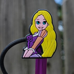 May include: A colorful charm featuring a cartoon image of a princess with long blonde hair, wearing a purple dress and tan sleeves. The charm is attached to a black band and a purple stick. The princess has blue eyes and a smiling expression.