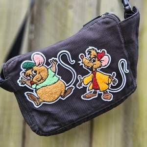 May include: A brown canvas messenger bag with two embroidered patches of cartoon mice. One mouse is wearing a green shirt and the other is wearing a yellow jacket with a red vest.