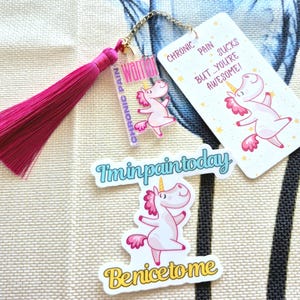Peut inclure: Un marque-page à pompon rose avec une chaîne dorée et un charm en acrylique transparent avec l'inscription "Chronic Pain Warrior" et un motif de licorne. Il y a aussi des autocollants avec des graphiques de licornes et le texte "I'm in pain today" et "Be nice to me."