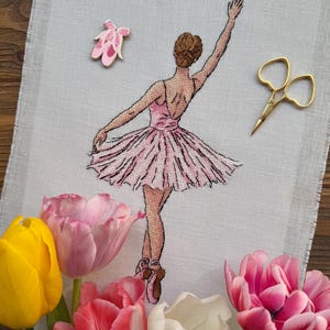 May include: A cross-stitch embroidery of a ballerina in a pink dress, with a matching ballet shoe pin and gold scissors. The image is decorated with colorful tulips in shades of pink, white, and yellow.