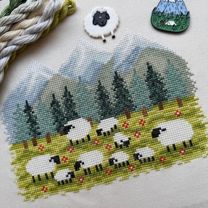 May include: Cross-stitch embroidery featuring a pastoral scene with sheep grazing in a green meadow, with a backdrop of mountains and pine trees. A small sheep-shaped pin and embroidery floss are also visible.