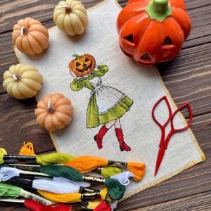 May include: A cross-stitch design of a woman with a pumpkin head, wearing a green dress and red boots. The image includes orange and yellow pumpkin-shaped candles, a ceramic jack-o'-lantern, red scissors, and embroidery floss in various colors.