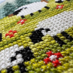May include: Close-up of a cross-stitch textile featuring a landscape scene. The design includes white, black, green, and yellow stitched areas, with small red flower accents. The stitching is dense and detailed, creating a textured appearance.