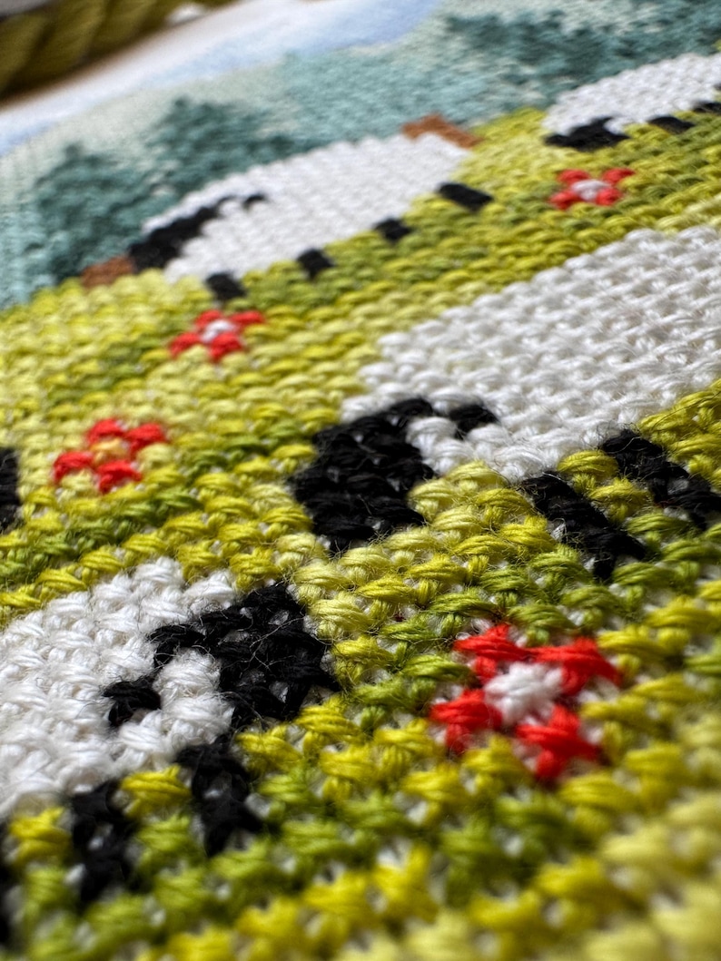 May include: Close-up of a cross-stitch textile featuring a landscape scene. The design includes white, black, green, and yellow stitched areas, with small red flower accents. The stitching is dense and detailed, creating a textured appearance.