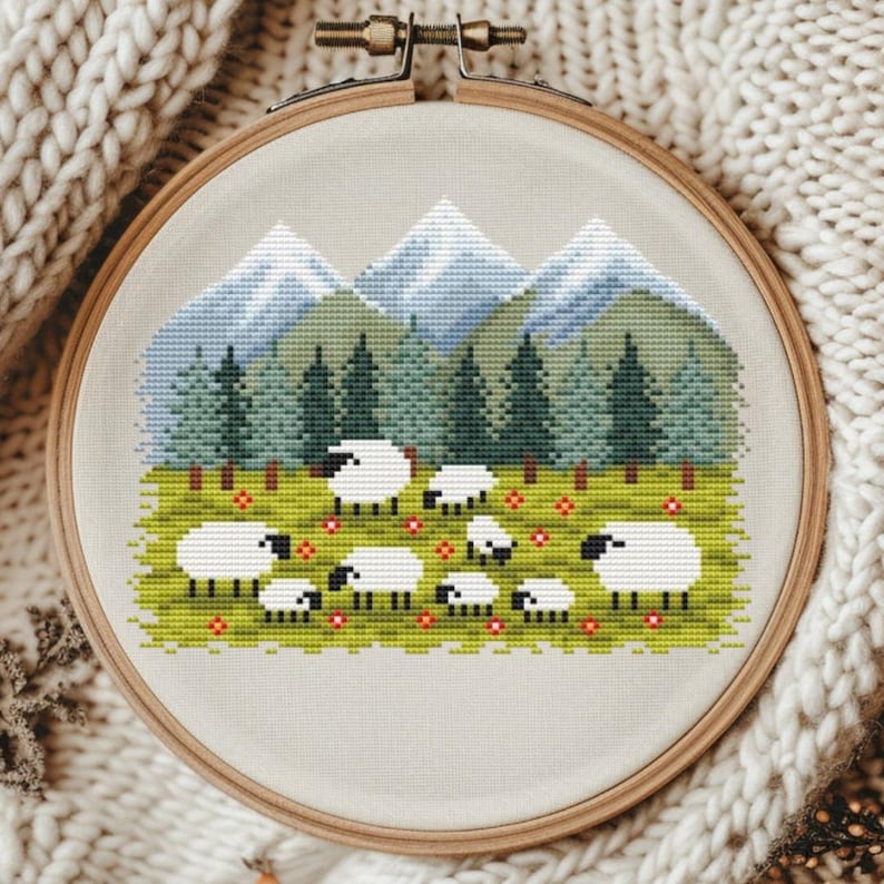 May include: A cross-stitch embroidery in a wooden hoop depicts a pastoral scene. White sheep graze in a green field dotted with red flowers, with a backdrop of green trees and blue mountains. The embroidery is set against a neutral fabric.