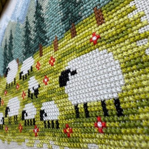 May include: A cross-stitch landscape featuring a flock of sheep grazing in a green meadow dotted with red flowers. The background shows a mountain range and evergreen trees. The sheep have white bodies and black faces and legs.