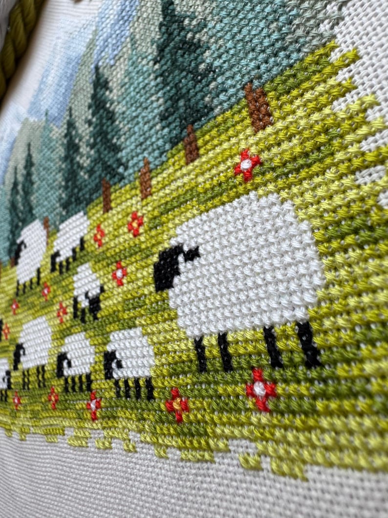 May include: A cross-stitch landscape featuring a flock of sheep grazing in a green meadow dotted with red flowers. The background shows a mountain range and evergreen trees. The sheep have white bodies and black faces and legs.