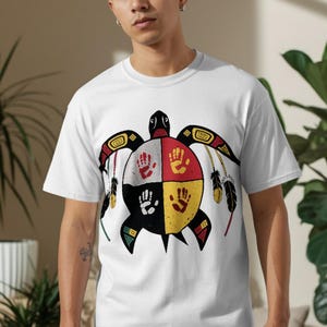 May include: White t-shirt featuring a turtle design with a Native American theme. The turtle's shell is divided into four sections, each with a handprint in red, yellow, black, and white. The design includes feathers and geometric patterns.
