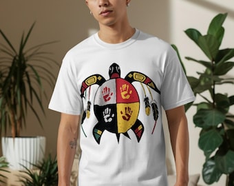 Può includere: T-shirt bianco con un disegno di tartaruga in stile nativo americano. Il guscio della tartaruga è diviso in quattro sezioni, ognuna con un'impronta di mano in rosso, giallo, nero e bianco. Il design include piume e motivi geometrici.