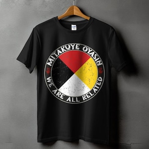 Op de afbeelding: Zwart t-shirt met een cirkelvormig ontwerp met rode, gele, zwarte en witte segmenten. De tekst "MITAKUYE OYASIN" staat er boven, met "WE ARE ALL RELATED" eronder. Het ontwerp bevat handafdrukken.