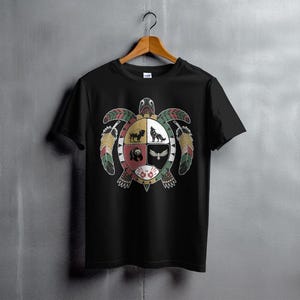 Puede incluir: Camiseta negra con un diseño de tortuga colorida con gráficos de animales y plumas. El diseño de la tortuga incluye un perro, un lobo, un oso y un búho. La camiseta está colgada en una percha de madera.
