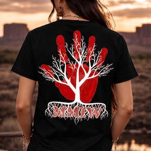 Op de afbeelding: Zwart t-shirt met een rood handafdrukontwerp, met een witte boomafbeelding en de tekst "MMIW" in het rood. Het ontwerp staat op de achterkant van het shirt. De achtergrond toont een zonsondergang boven een woestijnlandschap.