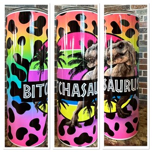 May include: A stainless steel tumbler with a pink, yellow, green, and blue ombre background. The tumbler has a black and white cow print pattern and features a graphic of a Tyrannosaurus Rex with the text "Bitchasaurus" in black.