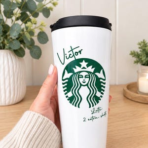 Könnte beinhalten: Ein weißer Reisebecher mit schwarzem Deckel und einem grünen kreisförmigen Logo einer Sirene. Der Schriftzug Victor steht über dem Logo, und der Text Latte 2 extra shots ist unter dem Logo in einer dunkelgrünen Schreibschrift gedruckt.