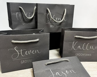May include: Several dark gray gift bags with silver rope handles. Each bag has a name in elegant script, with titles like "Groomsman" and "Best Man" below. The bags are arranged on a light wood surface.