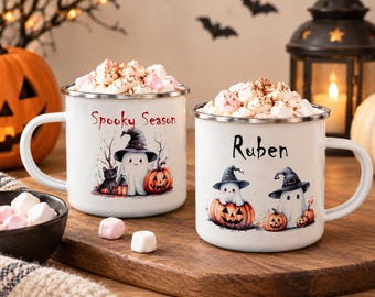 Könnte beinhalten: Zwei weiße Emaille-Tassen gefüllt mit Marshmallows. Die linke Tasse zeigt einen Geist mit Hexenhut, eine schwarze Katze und den Text Spooky Season. Die rechte Tasse zeigt zwei Geister mit Hexenhüten auf Kürbissen und den Namen Ruben.