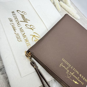May include: A brown leather zippered pouch with a gold zipper and a gold inscription that reads "Middleton-Keen family adventures" with a heart and arrow. The pouch is sitting on top of a white felt pouch with a gold inscription that reads "Emily & Ken Wedding Memories 6th October 2023".