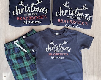 May include: A set of three navy blue t-shirts with the text "christmas with the Braybrook's" and the names "Mummy", "Daddy", and "Isla-Mae" in red. The shirts are folded on a wooden surface.