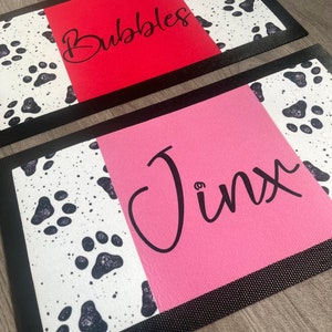 May include: Two black-rimmed doormats with a white background and black paw prints. The top mat has a red background with the name "Bubbles" written in black. The bottom mat has a pink background with the name "Jinx" written in black.
