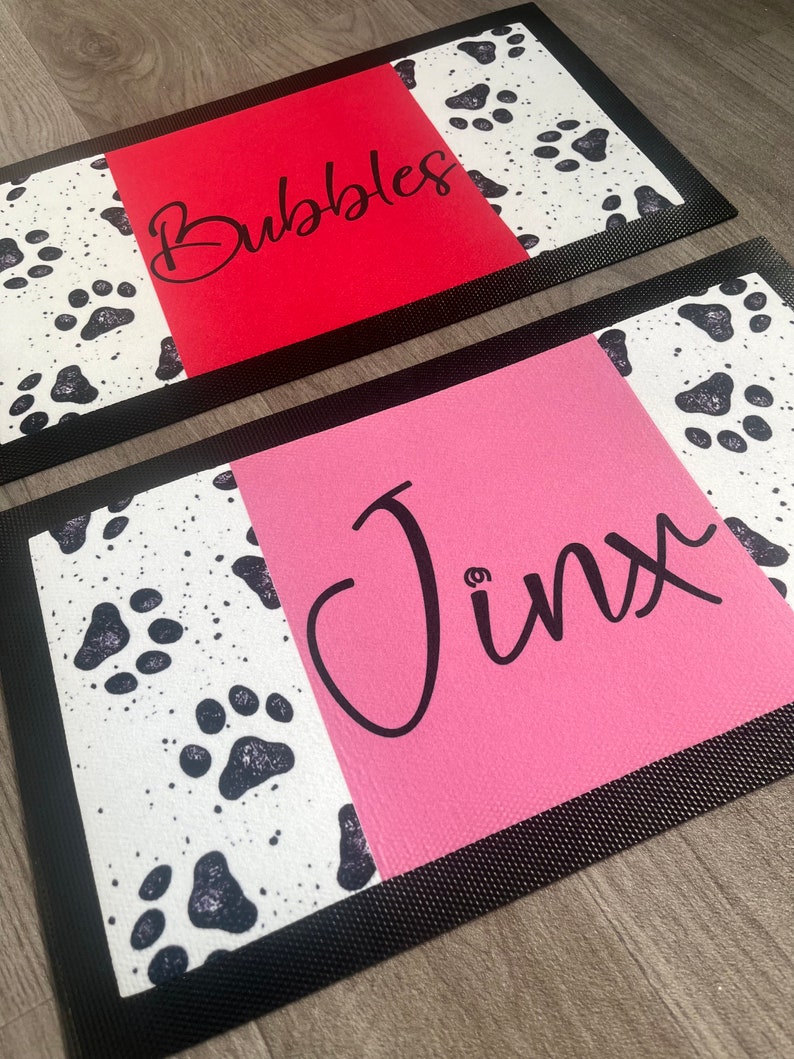 May include: Two black-rimmed doormats with a white background and black paw prints. The top mat has a red background with the name "Bubbles" written in black. The bottom mat has a pink background with the name "Jinx" written in black.