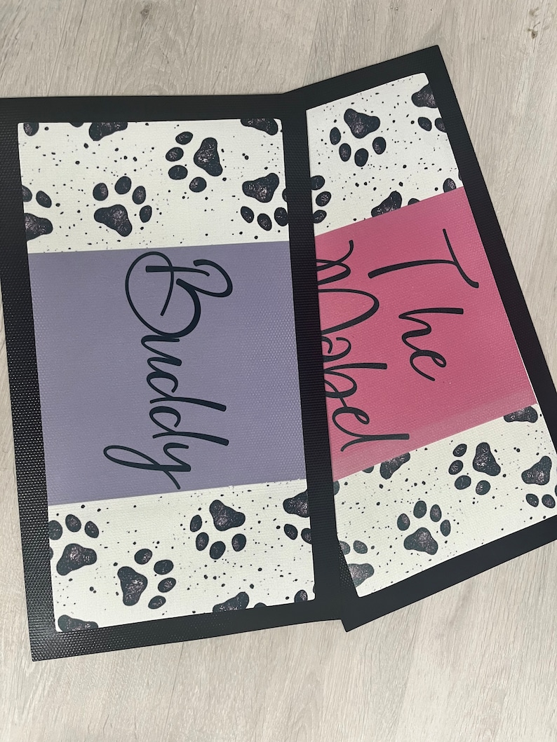 May include: Two black and white doormats with paw prints. The top mat has the text "The Rebel" in pink and the bottom mat has the text "Buddy" in purple.