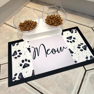 May include: White cat food bowl set with two bowls and a stand. The bowls are shaped like cat ears and contain brown cat food. The set is on a black and white paw print mat with the word "Meow" printed on it.