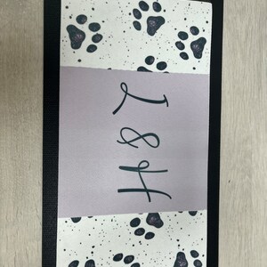 May include: A black and white doormat with a paw print design. The doormat has a purple section with the text "H&L" written in black.