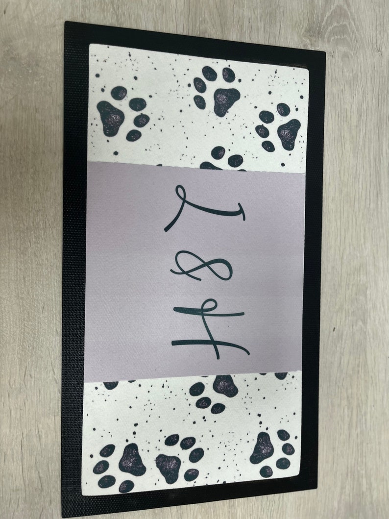 May include: A black and white doormat with a paw print design. The doormat has a purple section with the text "H&L" written in black.