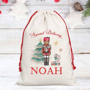 May include: A white canvas gift bag with red drawstrings. The bag features a printed design of a nutcracker standing next to a Christmas tree with a mouse holding a gift box. The text "Special Delivery" and "NOAH" are printed on the bag.