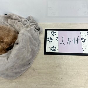 May include: A small brown dog curled up in a grey blanket, with a black dog lead attached to its collar. A black and white doormat with paw prints and the text "L & H" is on the floor.