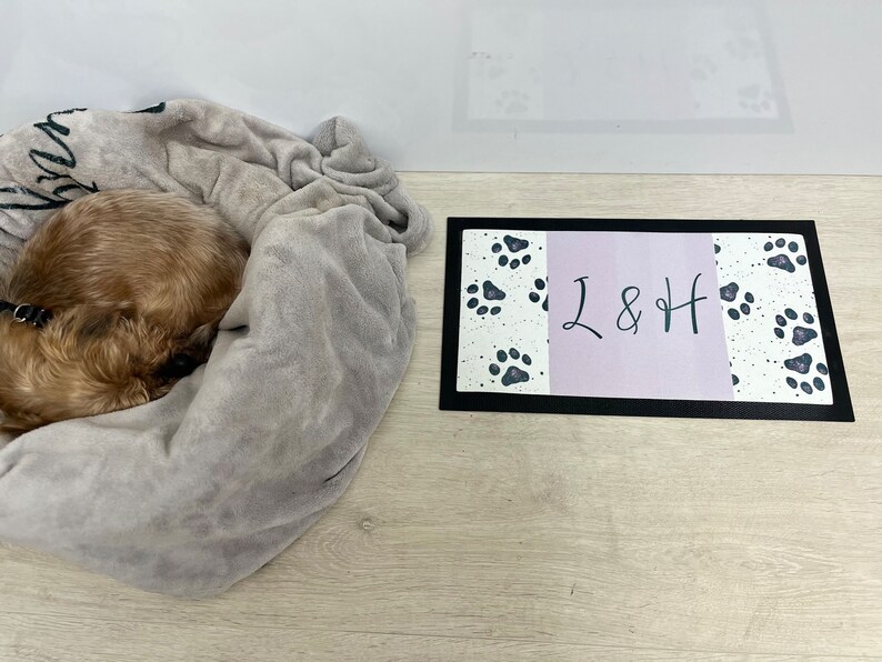 May include: A small brown dog curled up in a grey blanket, with a black dog lead attached to its collar. A black and white doormat with paw prints and the text "L & H" is on the floor.