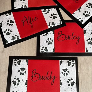 May include: Five red and black dog placemats with white paw prints. The placemats have black borders and the names "Alfie", "Buddy", "Bailey", "Harry", and "Lonnie" are written in black cursive on the red sections.