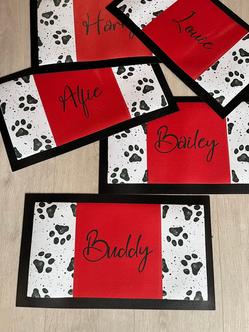 May include: Five red and black dog placemats with white paw prints. The placemats have black borders and the names "Alfie", "Buddy", "Bailey", "Harry", and "Lonnie" are written in black cursive on the red sections.