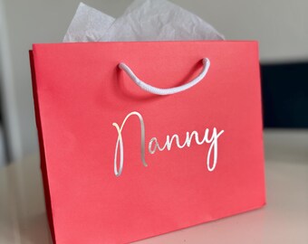 May include: A bright pink gift bag with white handles and the word "Nanny" printed in silver foil.